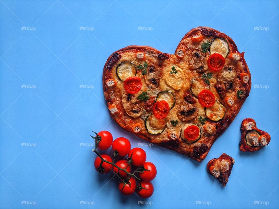 Delicious vegetarian pizza in the shape of a heart for Valentine's Day with tomatoes, vegetables and cheese on a blue background.