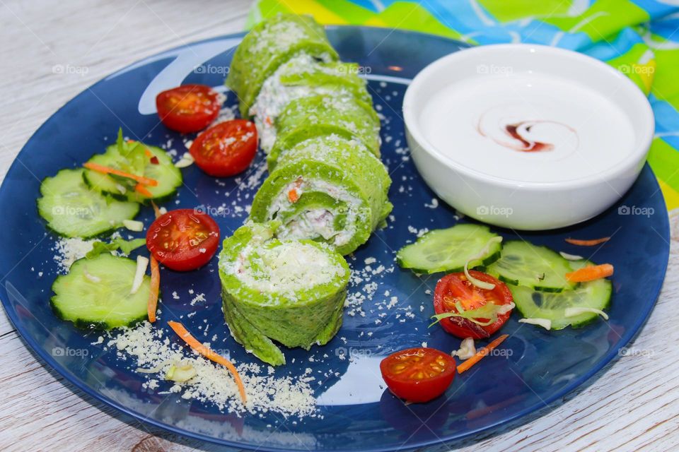 Rolled spinach pancakes filled with cream cheese and vegetables on a blue plate with cherry tomato and yogurt