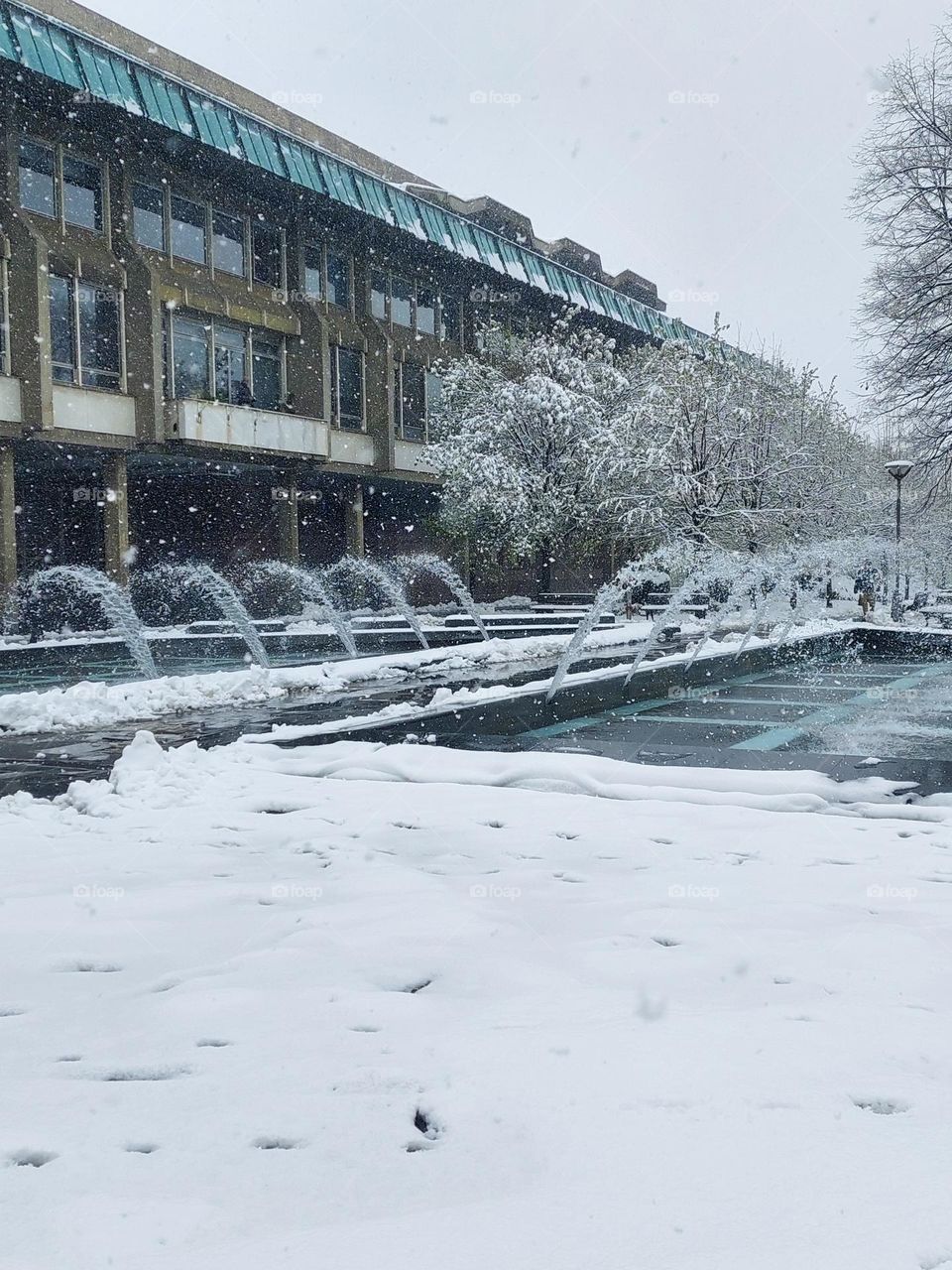 A city park with a fountain covered in snow.  It's snowing