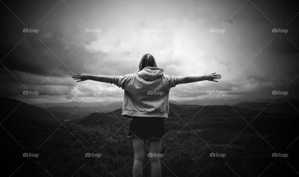 Black and white picture of a young woman with arms open wide in front of mountain range.