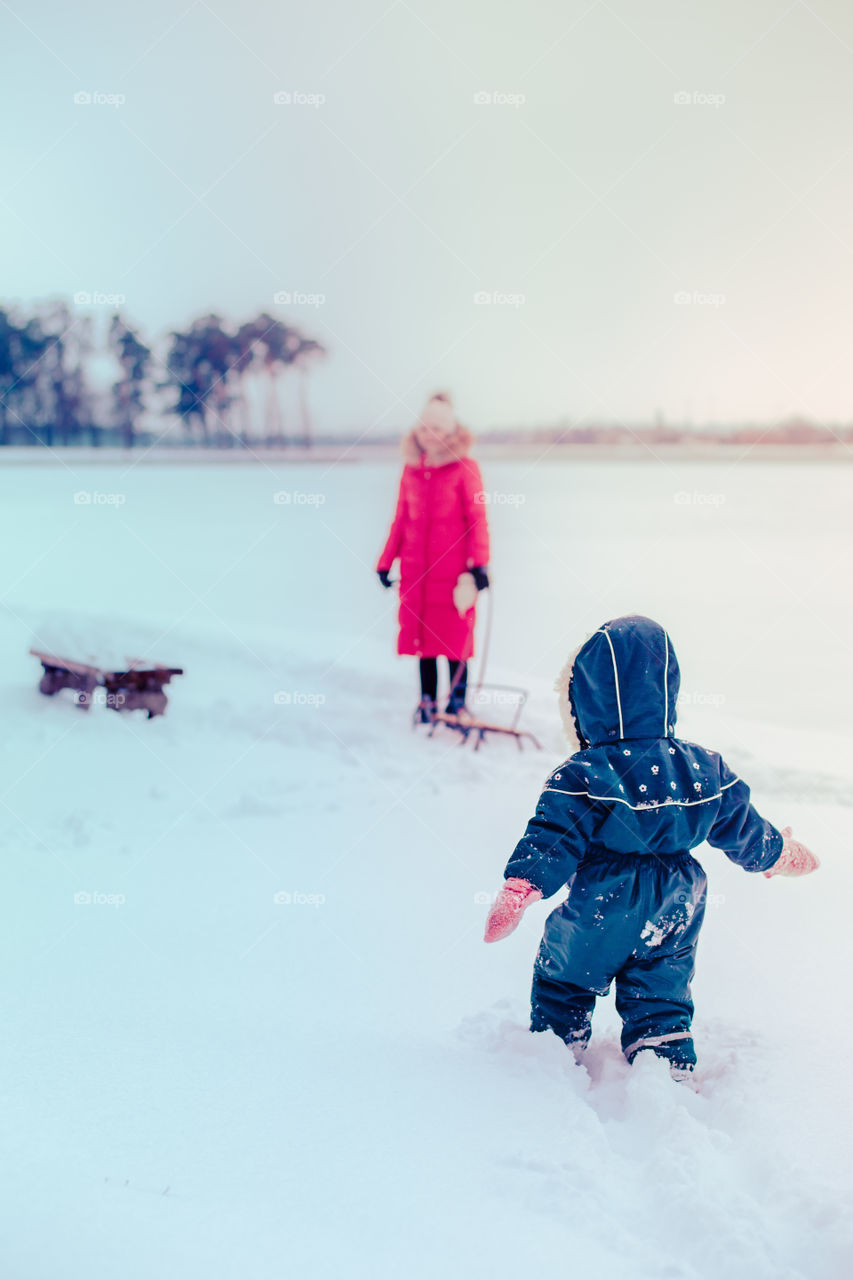 Mother and her little daughter are spending time together walking outdoors in forest in winter while snow falling. Woman is pulling sled, a few years old girl is walking through the deep snow, enjoying wintertime