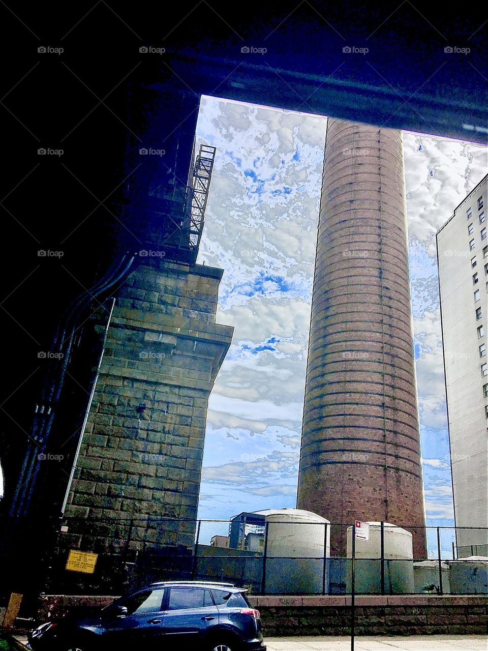 An unusual strange angle looking up at the “Manhattan Bridge” in downtown NYC. The skyscraper shows the bandwidth of the dimensions. Monumental architecture dwarfs by comparison the pickup trucks driving by.