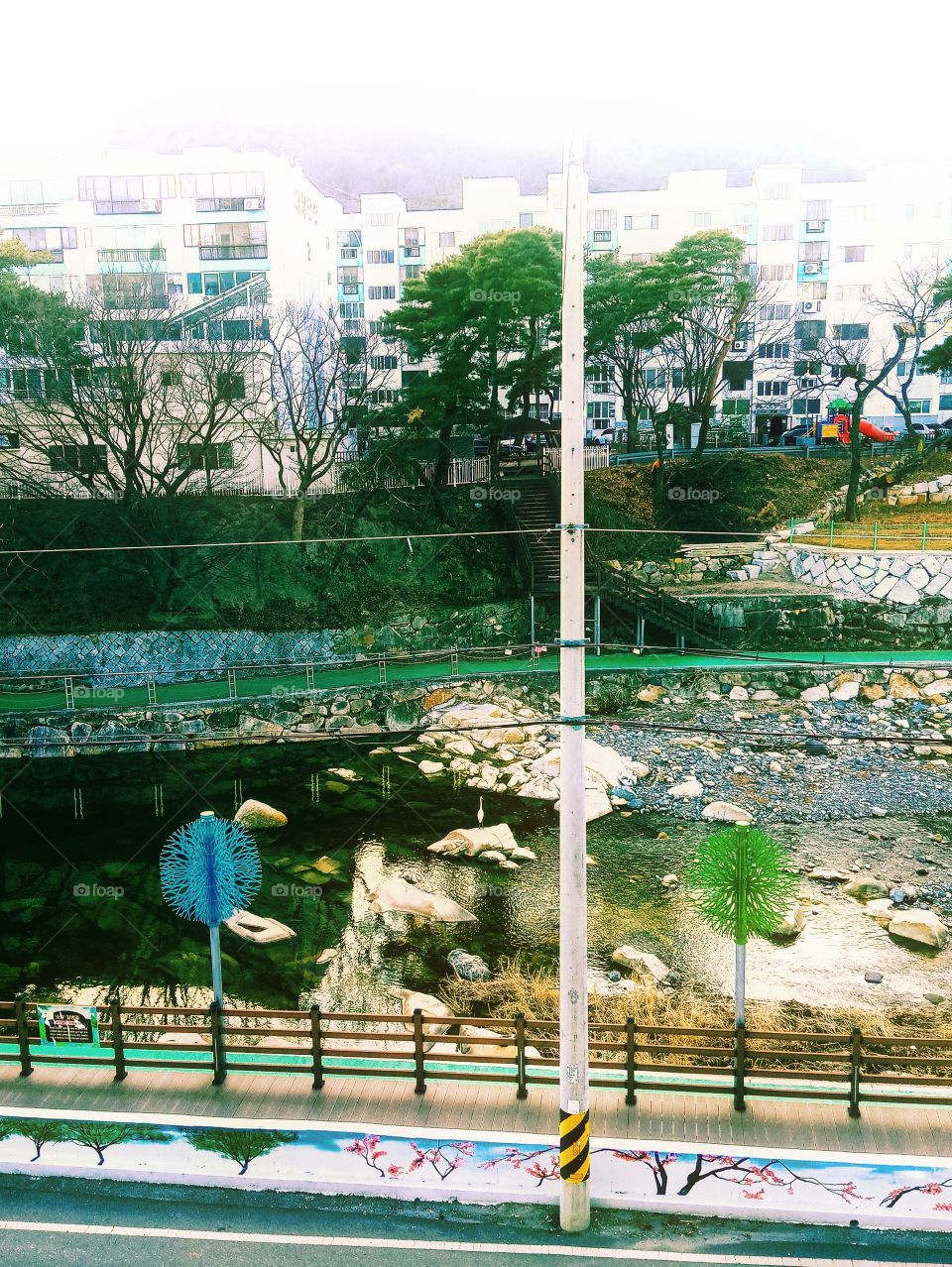 The River Bank in front of our home w/ the big king fisher on top of the big rock in the middle of the river , the green passage for the people to walk on beside the river, the stairs going up, the building in front of the mountains.
