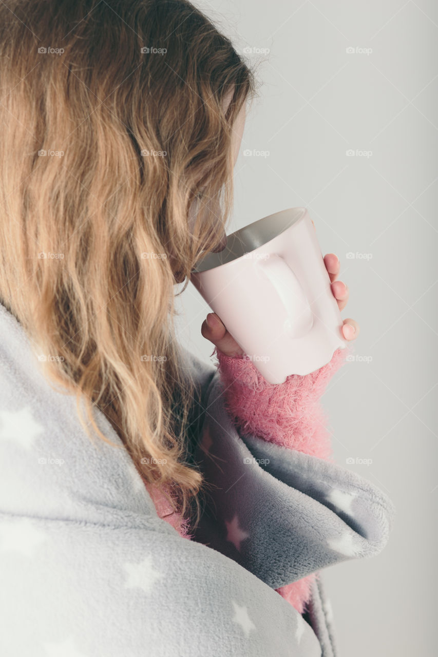 Young woman has caught cold, has day off, sick day, wrapped in blanket, holds cup of herbal tea. Profile portrait view