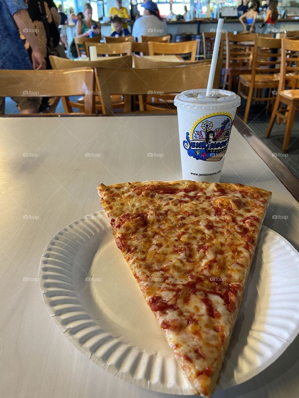 A pizza slice and soda from a restaurant counter at the boardwalk. What could be more authentic for Jersey Shore summer meal. Everything tastes better on the boardwalk. People in the background are enjoying seafood, sushi, and drinks at the bar. 