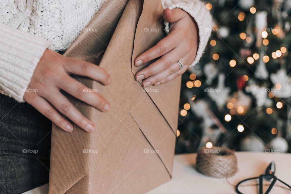 Midsection of woman wrapping christmas present in brown paper in front of christmas tree at home