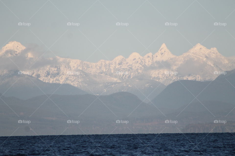 Shot of the Northwest Pacific Coast glowing with the rare winter sunlight. The sea is indigo blue, the hills behind dark green, mountains capped with fresh snow and small wisps of powdery clouds kiss the mountain peaks.