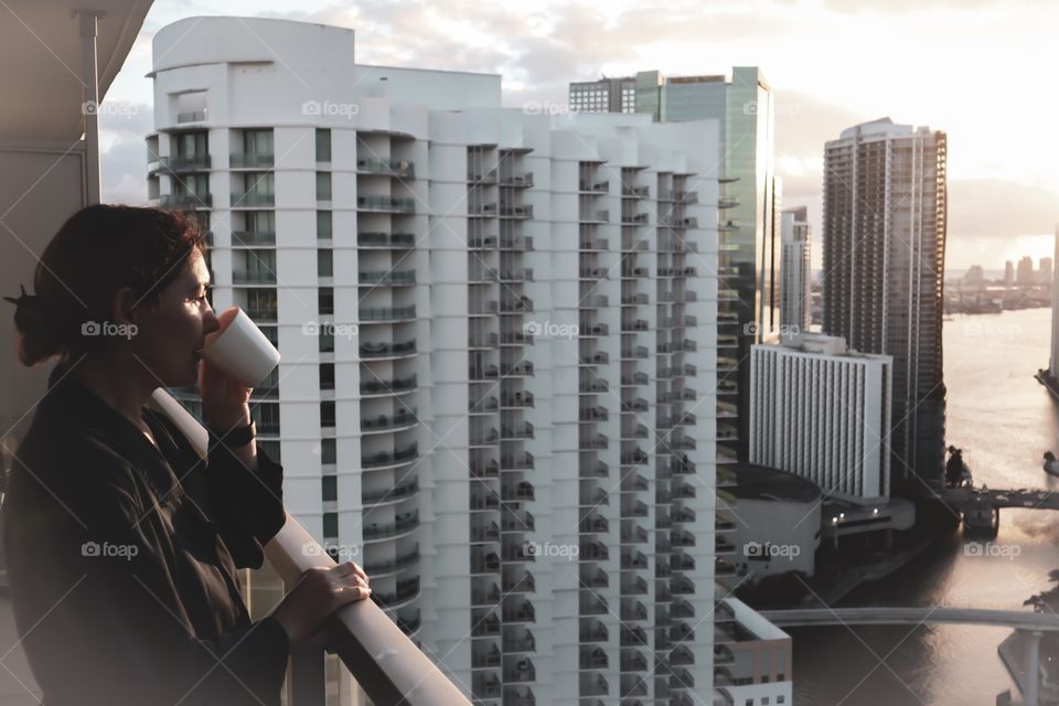 woman in bathrobe drinking Her morning coffee or tea on a downtown balcony. beautiful sunrise in downtown Miami. Woman enjoying Miami downtown city view from a balcony. 