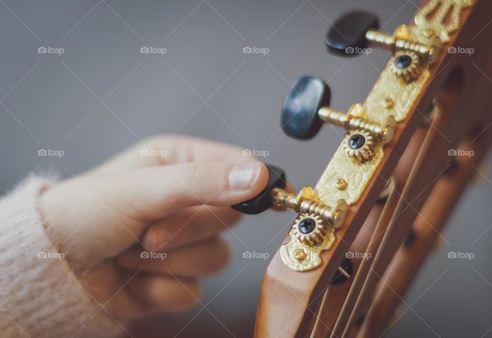 One hand of a little caucasian girl holds a guitar and with her fingers clamps a peg while adjusting the strings on a guitar socket while sitting in a room on a sofa, side view close-up. Music education concept.