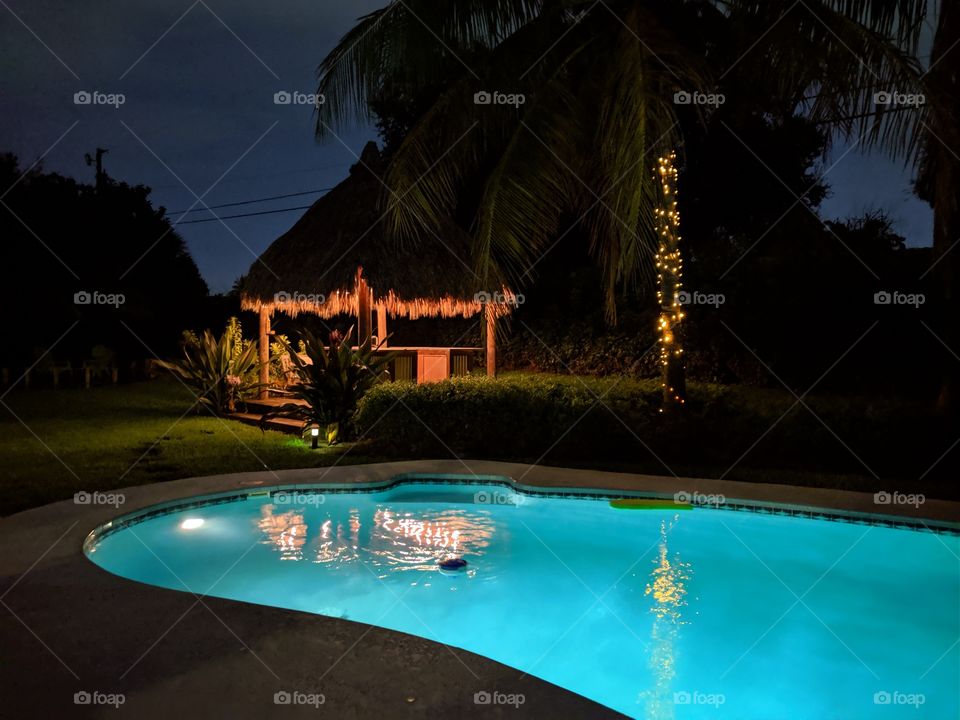 Nighttime image of an illuminated pool and tiki hut next to a palm tree