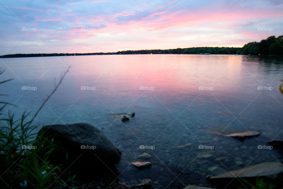 Beautiful pink summer sky reflection on clear water lake at sunset with rocks and pebbles 