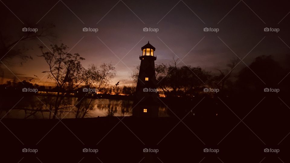 Lighthouse is standing by. Sky is multicolored with layers of high clouds not letting good light through. Reflections of shore line can be seen mirrored on Lake Waters.