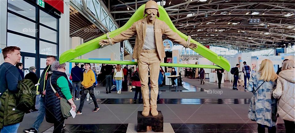 Is it a contraption for a circus act, some kind of projectile for a flying man or simply an oversized green clothes hanger he is attached to ? His goggles brought on the flight idea. It was seen at a trade show in “Munich”. 2024. Hypnotic Productions