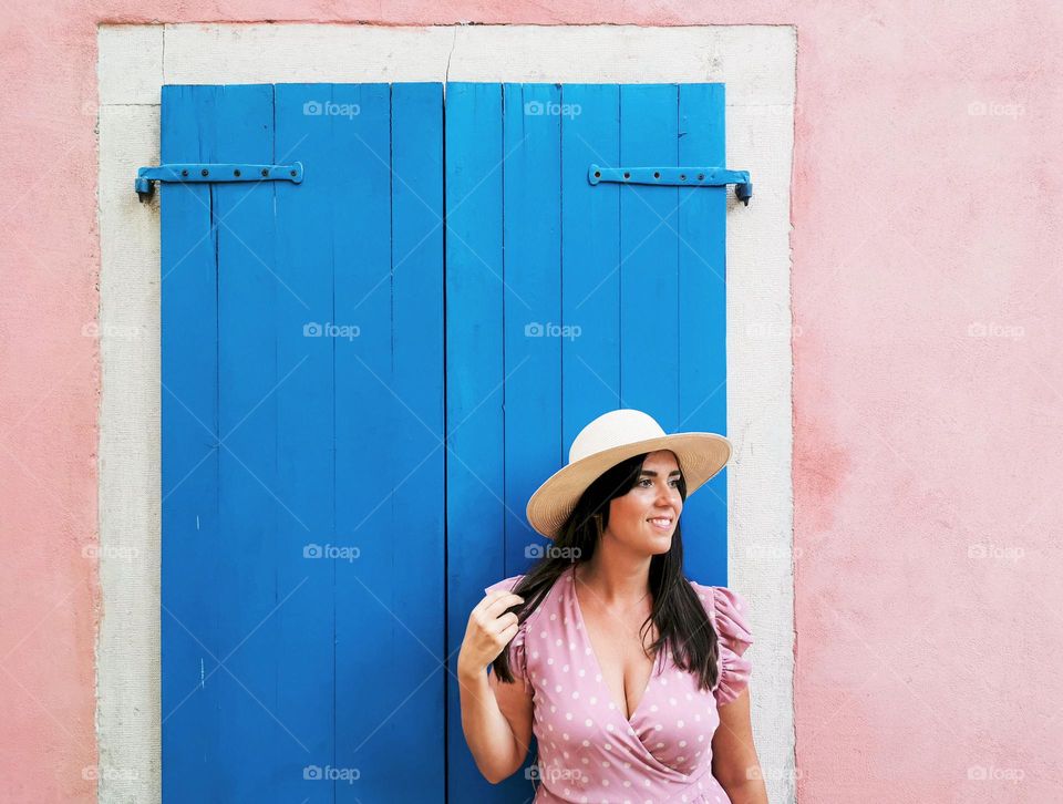 Portrait of young woman wearing stylish summer clothes and sun hat, standing in front of window with blue shutters