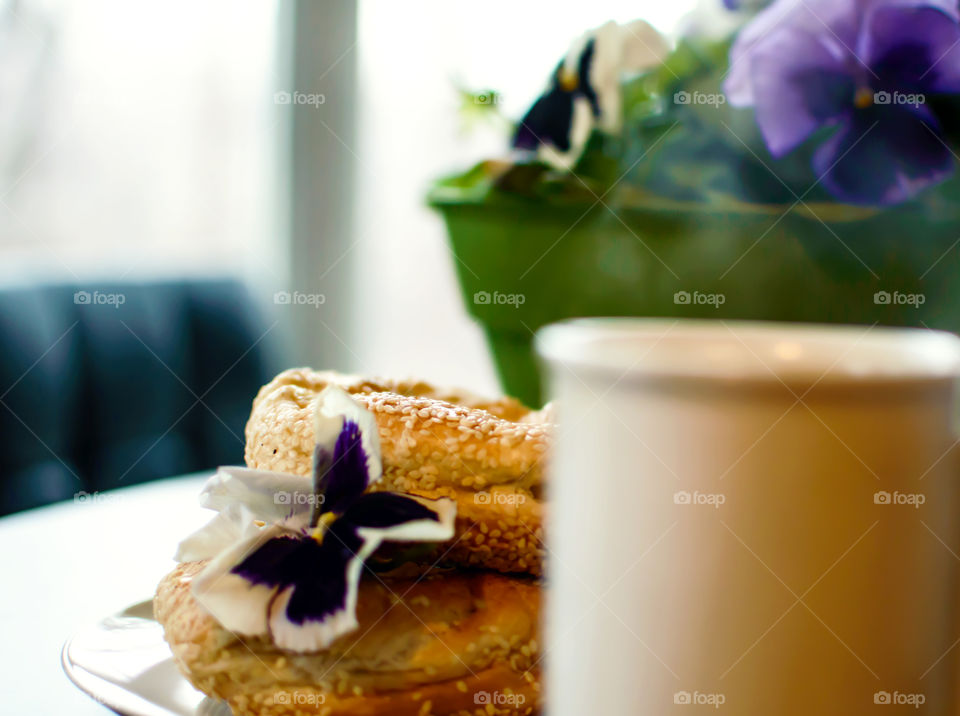 Steaming cup of tea or coffee with fresh bagels on tabletop decorated with pansy flowers in morning for light breakfast background 