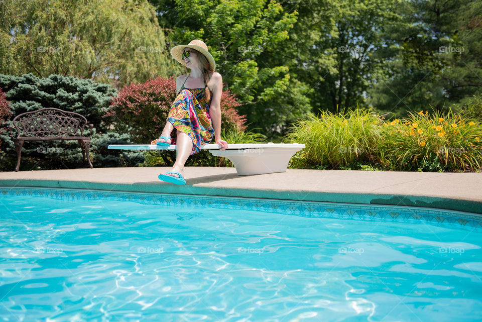 Young millennial woman wearing a dress and sunhat while sitting on the diving board of a swimming pool in the summer