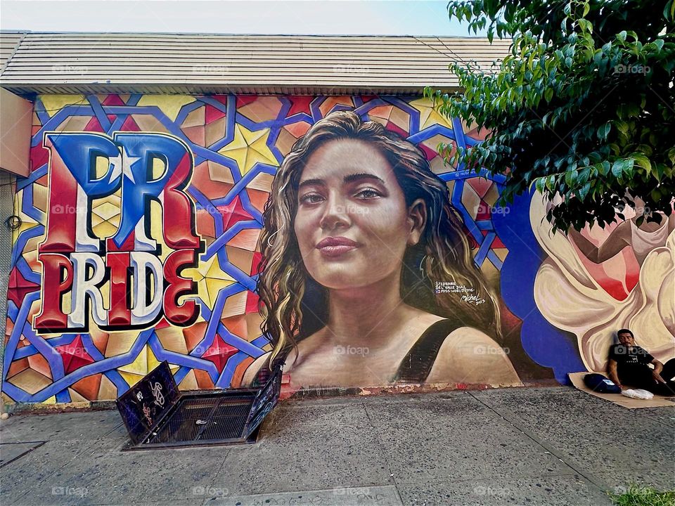 This smiling Latina next to the words “PR” and “PRIDE” is part of a large art mural filling almost an entire city block at “Jefferson St” and “Broadway” in “Bushwick”, Brooklyn celebrating the Latin culture. 2024. Hypnotic Productions