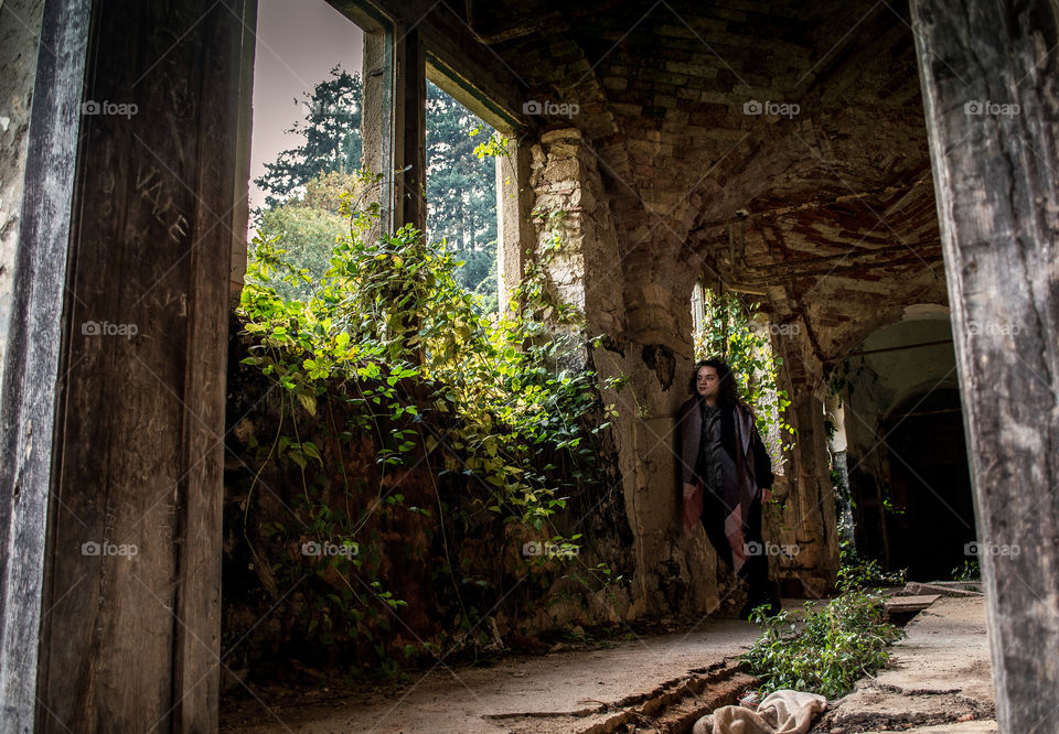 Curly girl standing next to the window covered with plants and greenery in an abandoned castle