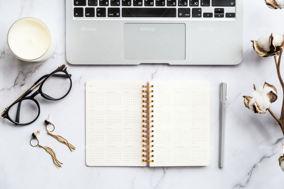 Desktop flatlay items: laptop, calendar, eyeglasses, candle, earrings, pen, cotton flowers lying on marble background 