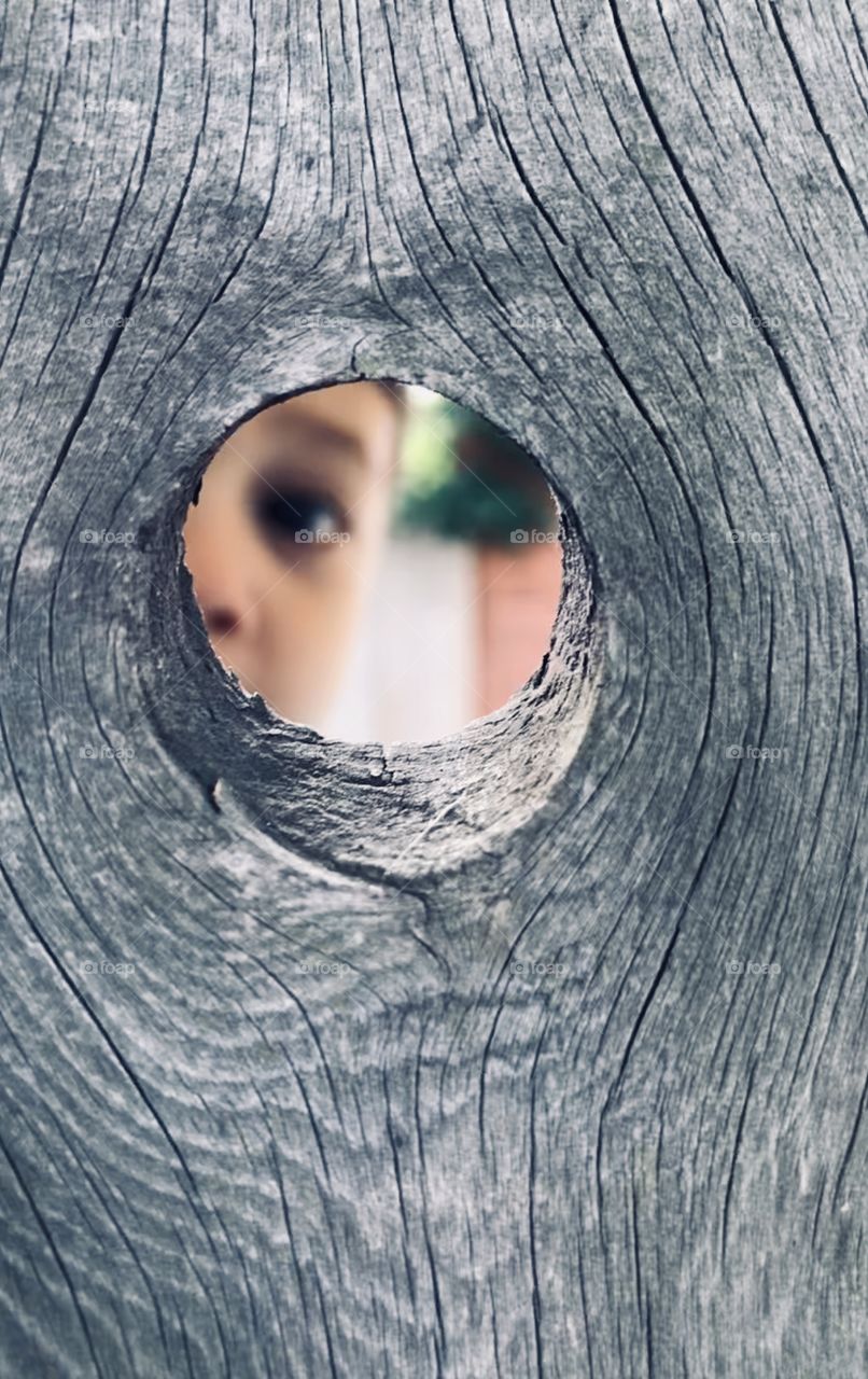 Young boy looking through a hole in a wooden fence