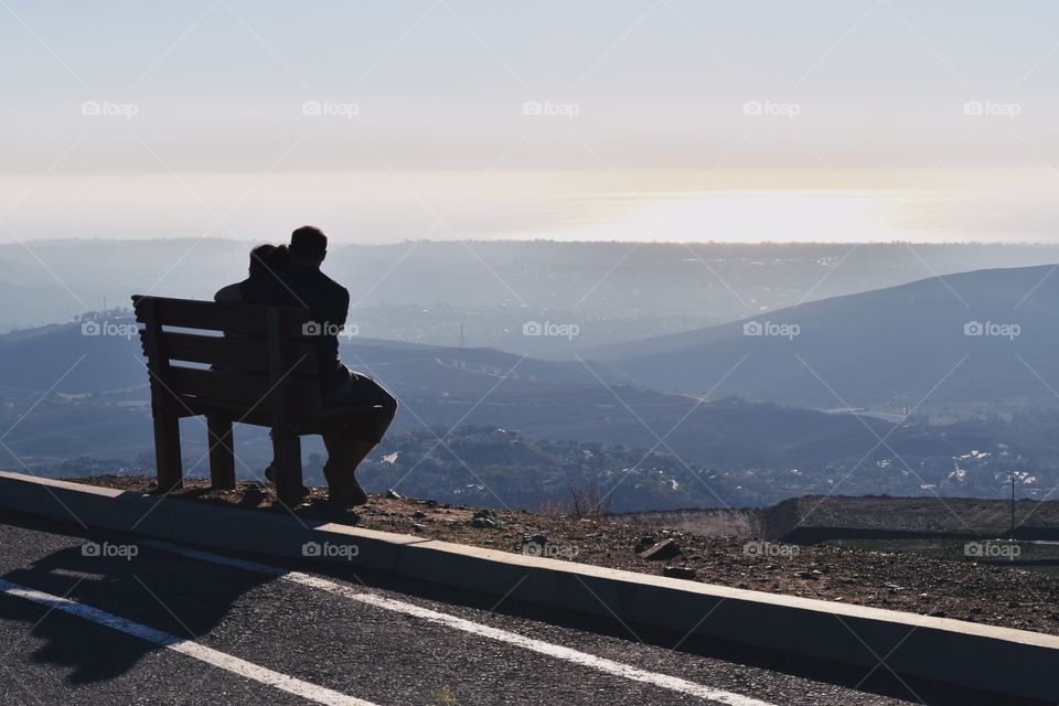 A couple, man and woman, are sitting on a bench on the top of a hill hugging enjoying view of a ocean