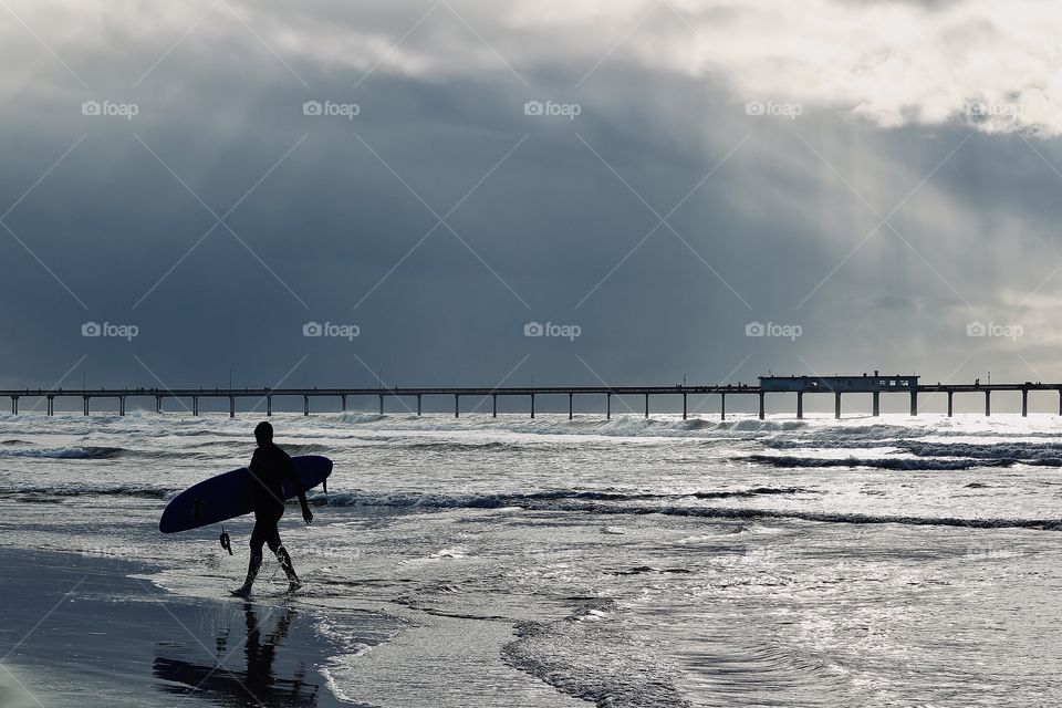 A male surfer emerges from the ocean waves in California. Summer rain clouds loom in the background as rays of sunlight push through.