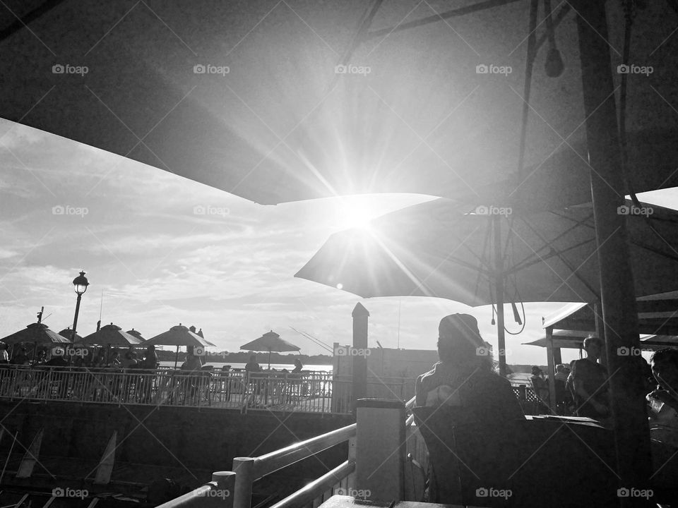 A black and white image of people dining at a bar/restaurant on the 9th Avenue Pier in Belmar, NJ. It’s a very relaxed place to eat and have a cocktail while looking at the water and the boats.  The sun shines down through the umbrellas. 