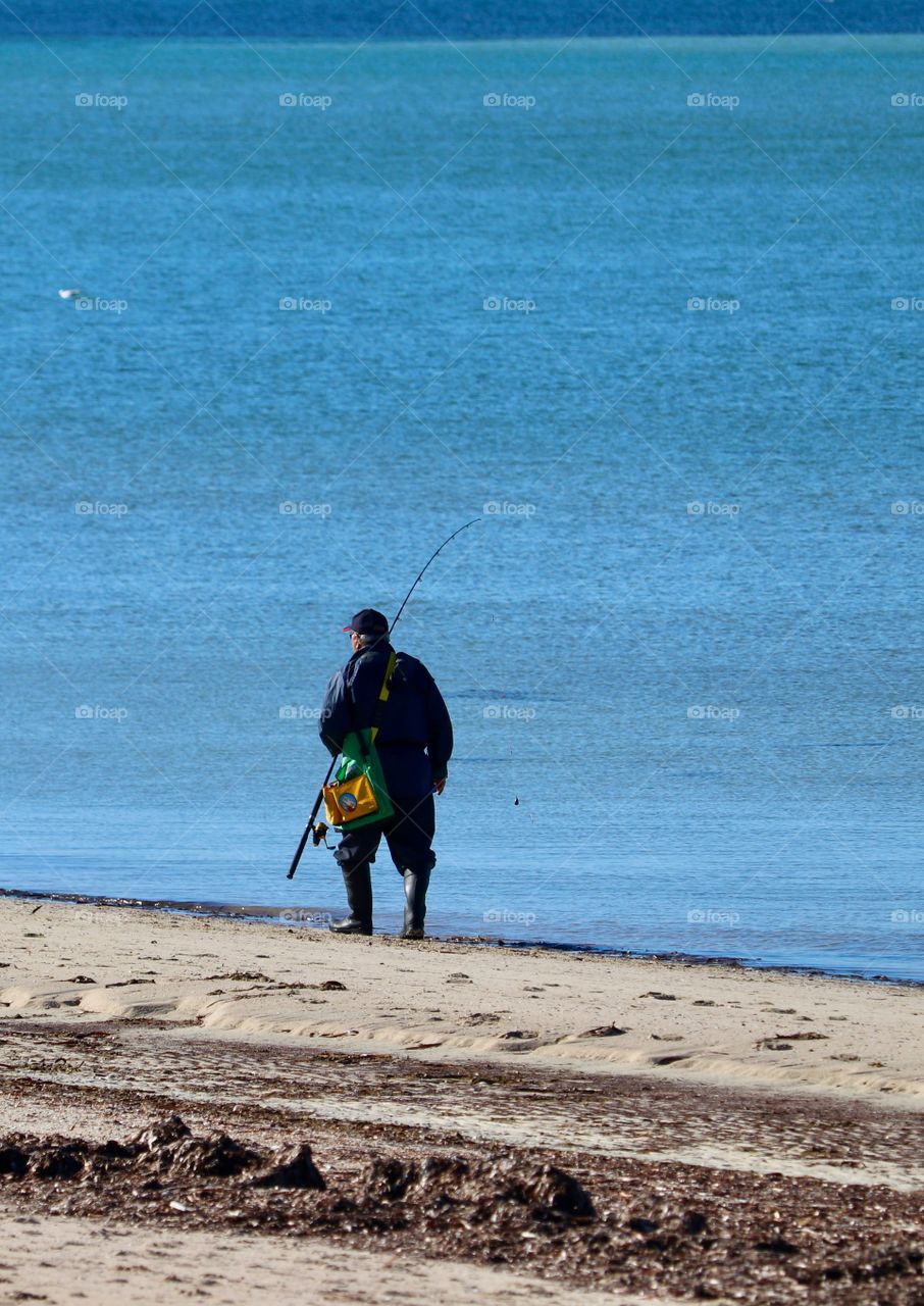 Lone fisherman in waders with equipment on beach at seashore south Australia 