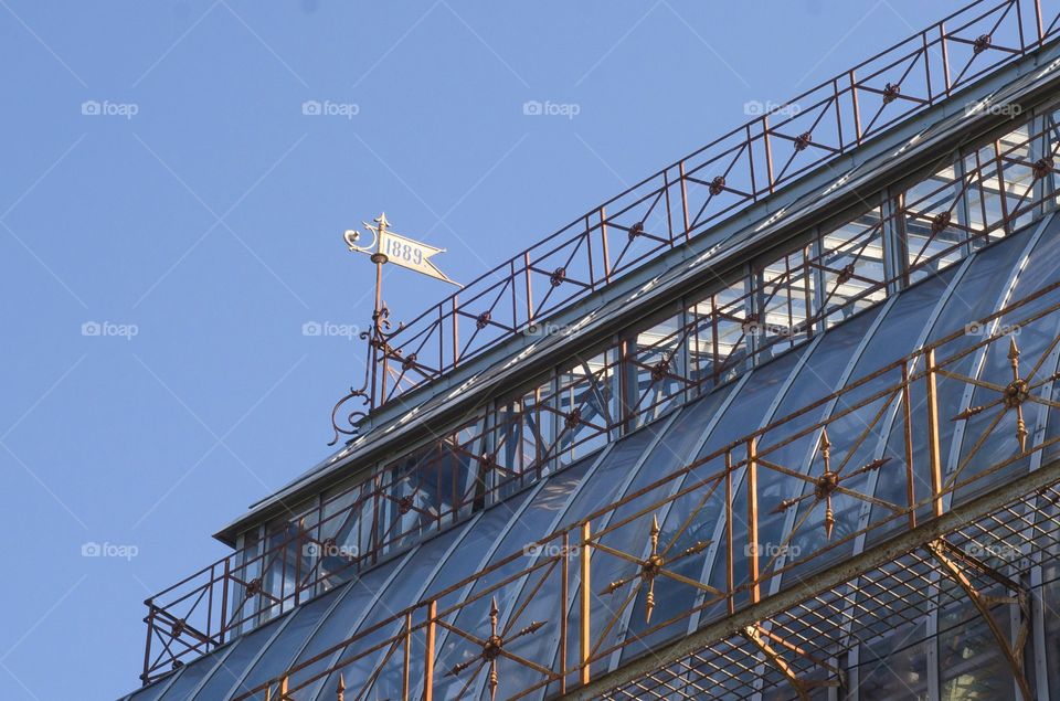 The roof of the greenhouse of the botanical garden. A metal flag with the designation of the year of construction. The structure is made of glass and metal. Elegant light design. Clear blue sky.