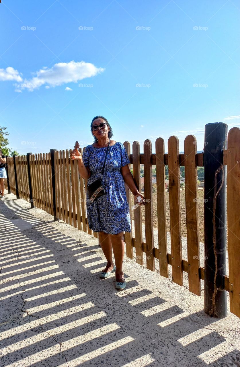 Senhora bem tranquila e natural num passeio: usando vestido azul com detalhes branco e preto, sapatilha azul claro, bolsa pequena preta,ócultos escuros e, com uma paleta mexicana na mão