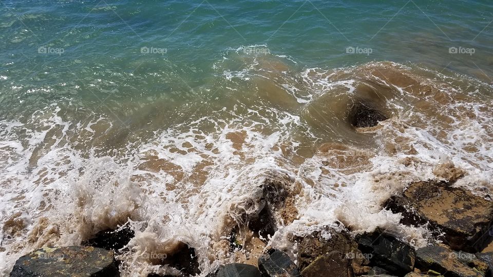 action shot of waves crashing against a rocky coastline mixing up sand and water. the water color fades from a deep sea green in the background to sandy brown foreground with seafoam garnish