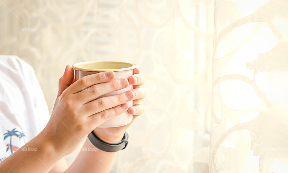 Hands hold a cup with beverage near the window. One person stay home and take care.