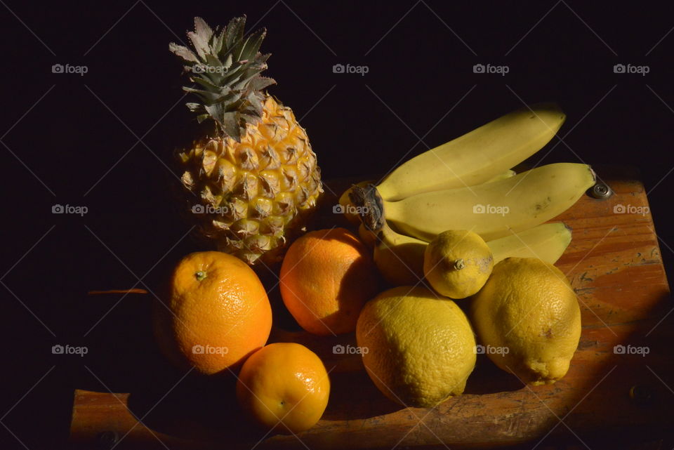 An arrangement of fruits. Pineapple, bannanas, oranges, lemons and a naartjie, all standing on a wooden stool.