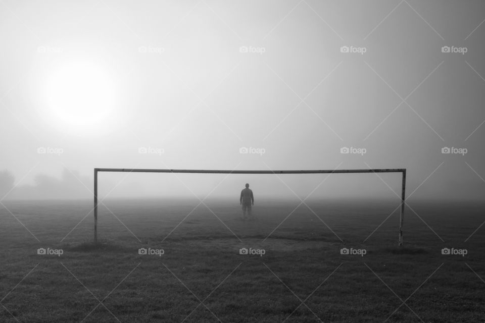 A lone footballer standing in the goal on a cold and foggy morning with no other players in sight.