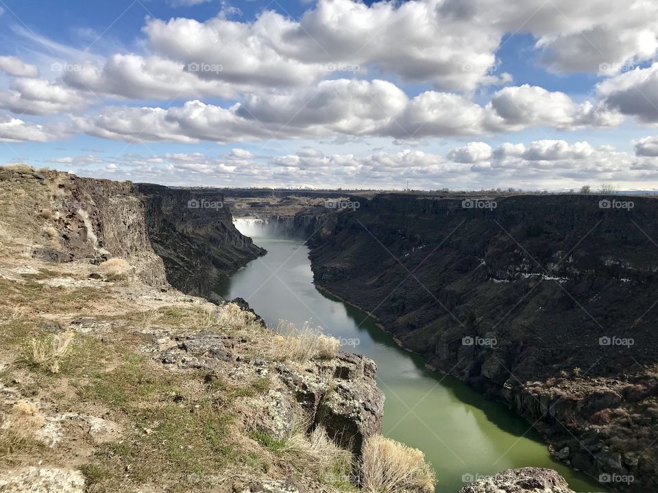 A breath taking view of the snake river canyon. The crisp blue partly cloudy sky casts shadows along one wall. And the sun shines down on the other side. In the center of the Canyon is the view of the Shoshone Falls.