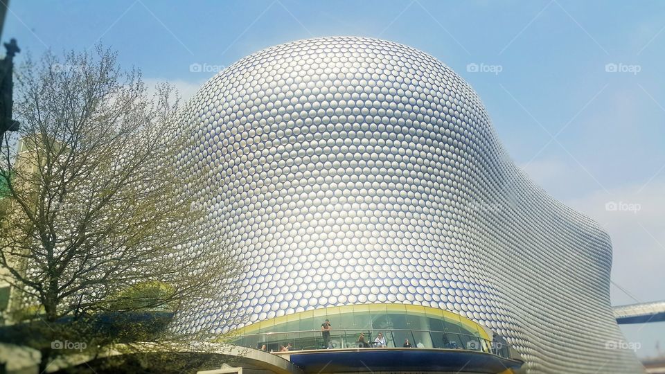 BLOBITECTURE - Selfridges Building. Composition - The anodized aluminium disc facade (patterns) is complemented by mild colours of the blue sky, green tree leaves, dark blue base,  yellowish-green arch above the glassy base area.