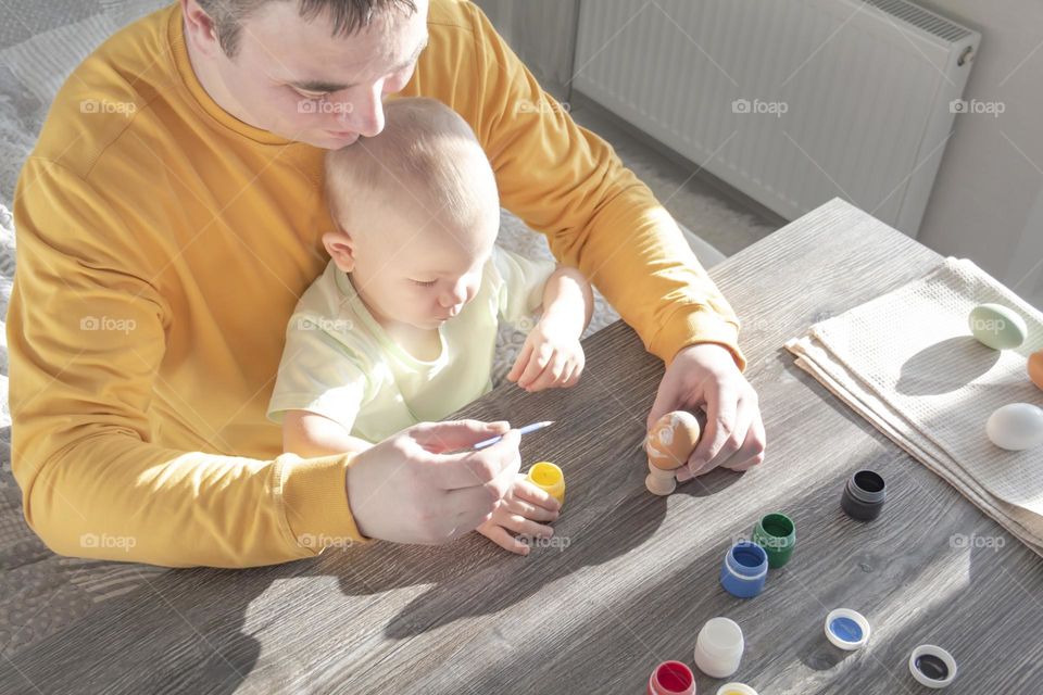 A little son and his father together in their free time paint Easter, chicken eggs with paints and brushes for the holiday. A child and a man create a craft and natural product in their family business.