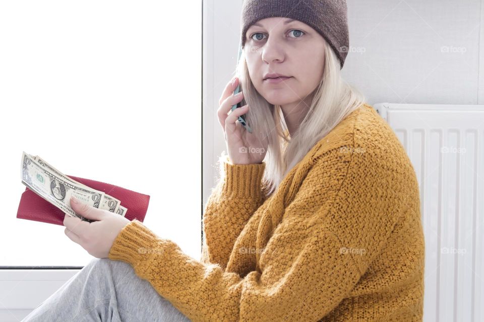 A young girl in a yellow sweater and a brown hat is sitting on the floor, counting money and thinking how to pay bills and taxes near a heater with a thermostat..