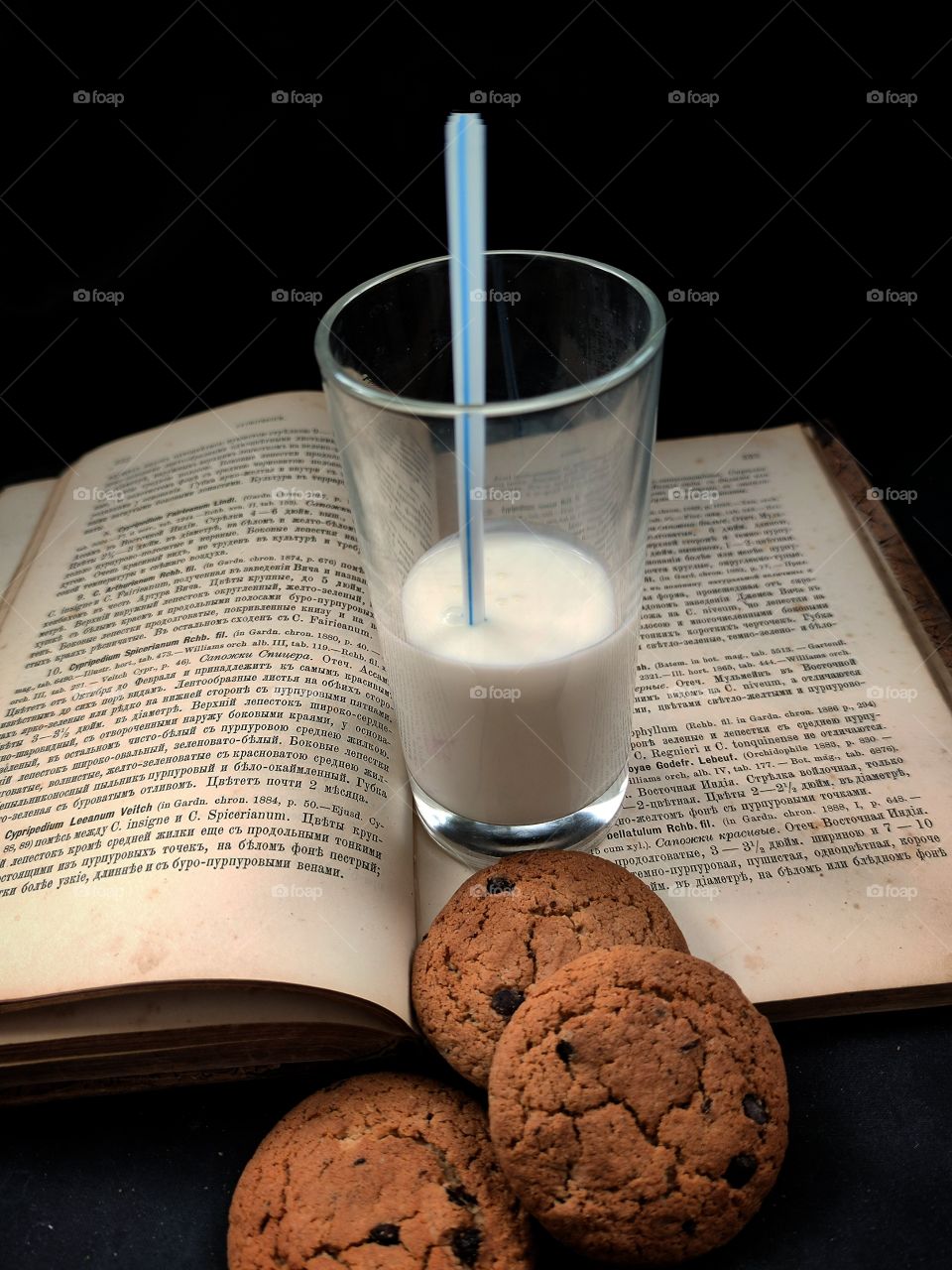 an old book with a glass of white milk and a blue straw and oatmeal cookies with a chocolate.  Black background