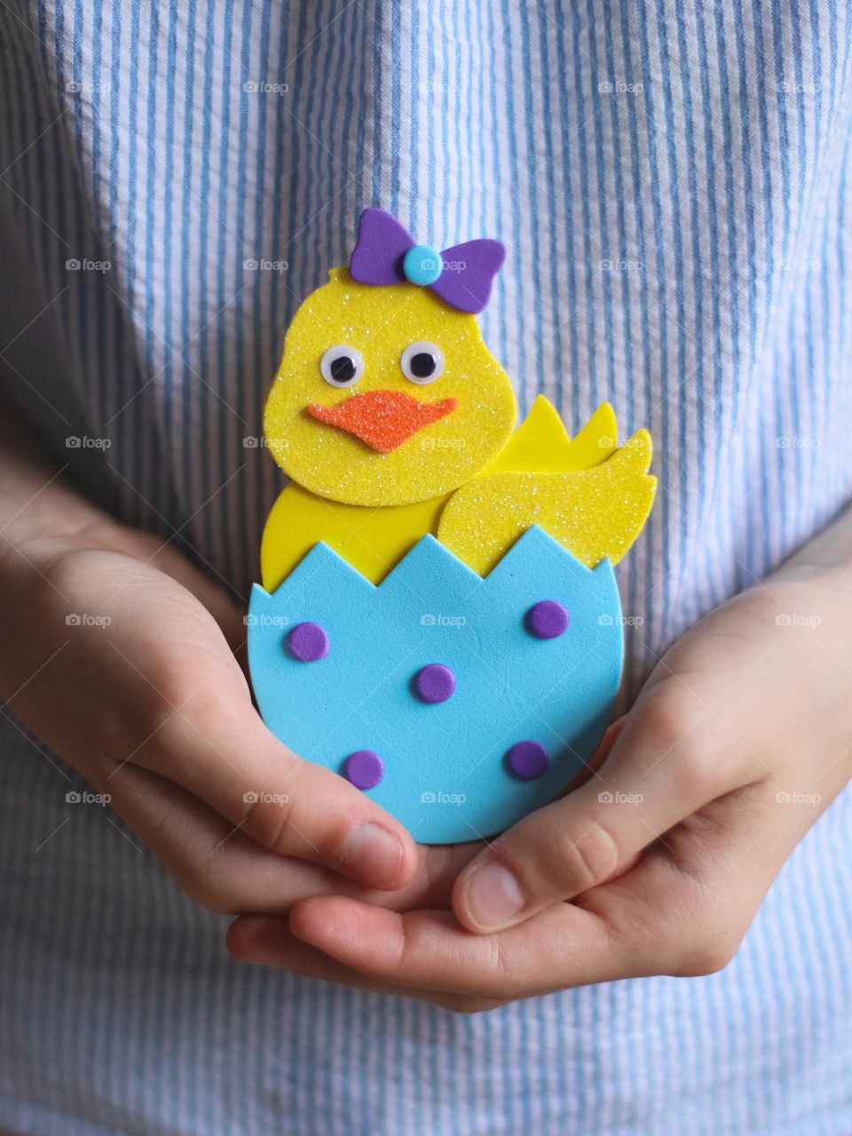 Hands of a caucasian teenager girl in a blue striped blouse are holding a homemade felt Easter chicken, close-up side view. Concept of creative kids preparing for Easter.