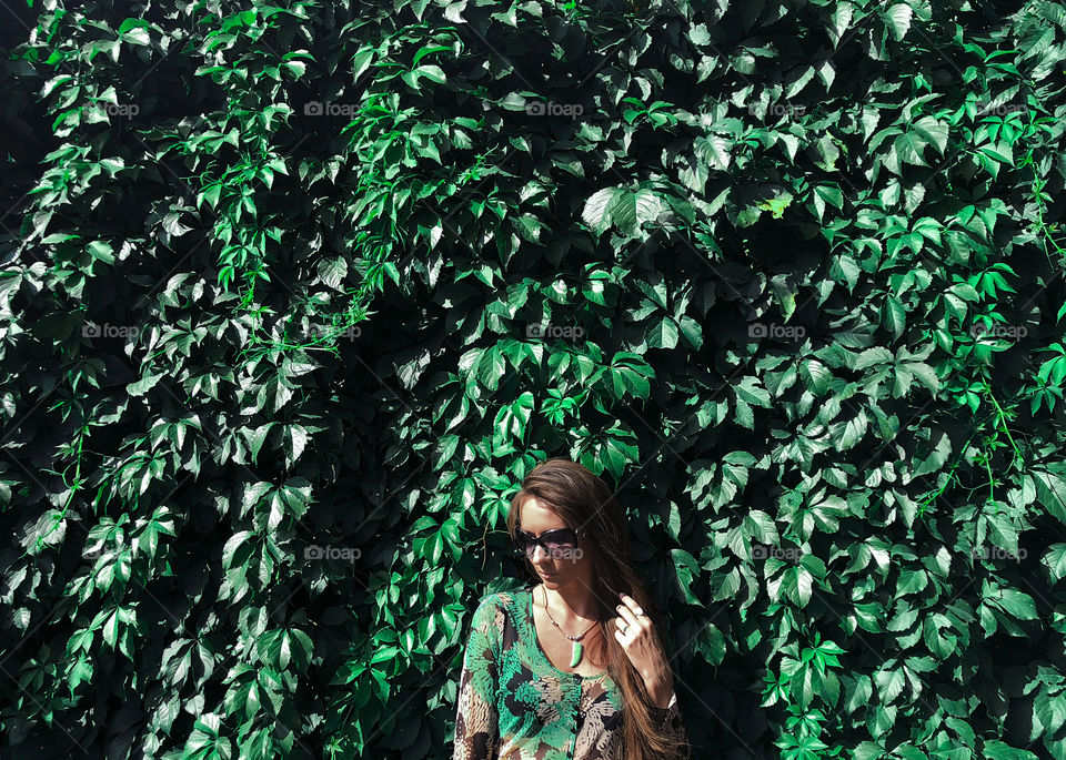 Young woman with long hair wearing green clothes standing in front of a green leaf wall 