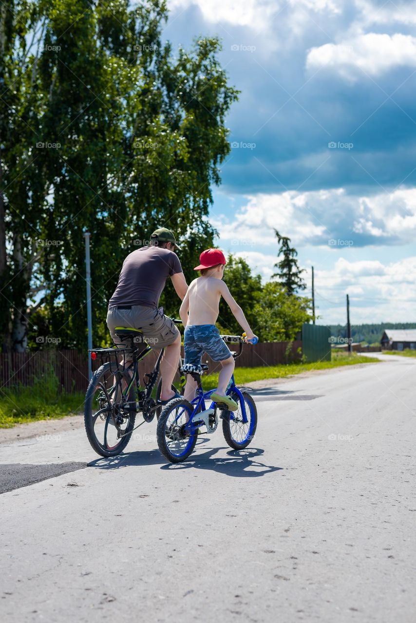 Father and son ride a bike down the street in the summer in the village