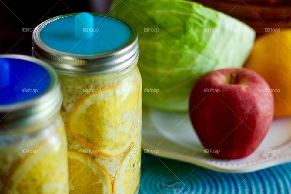 Fermented cabbage, apples and oranges in quart-size mason jars with blue silicone fermenting lids, a cabbage, an apple, and an orange on a white plate on a blue placemat in the background 