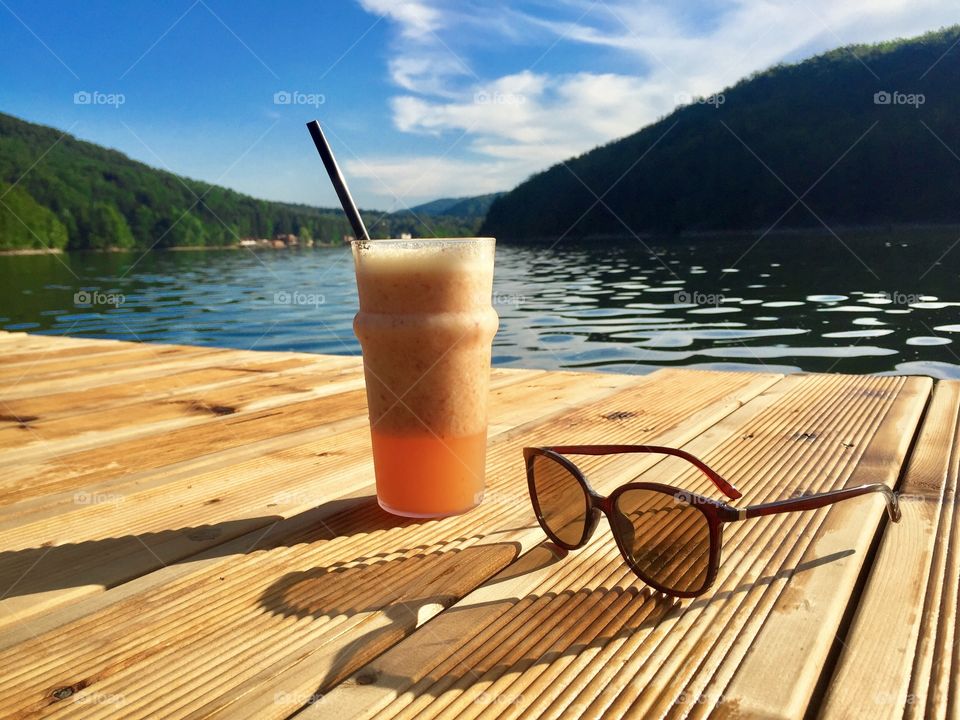 Glass of orange smoothie with drinking straw with a pair of sunglasses on a rustic wooden pontoon near the lake