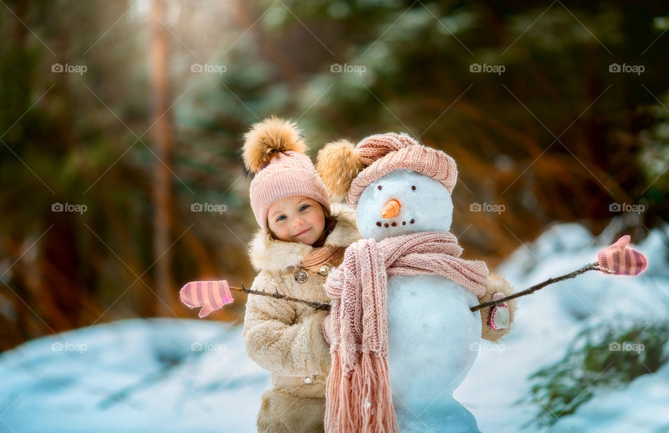 Little girl with snowman in winter forest at sunny day