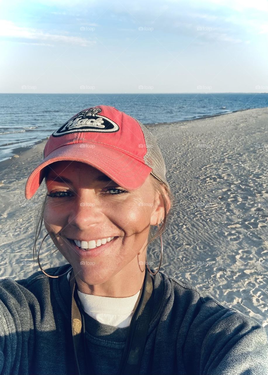 Happy Woman smiling wearing a red trucker hat on sandy Lake Eerie beach with lake behind her