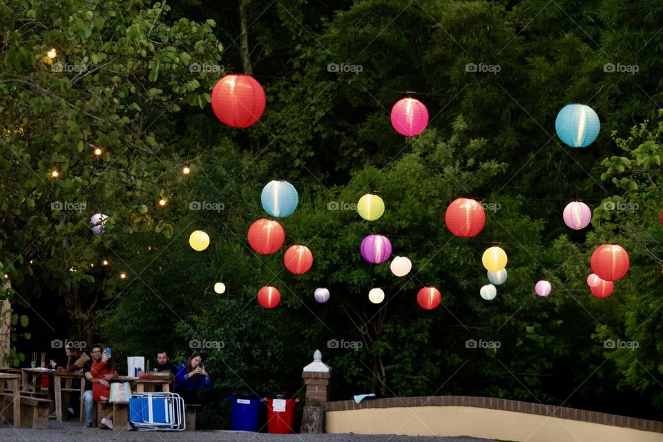 Bright and colorful lanterns suspended in the air illuminate a lush forest scenery, creating a magical environment. Picnic tables are visible in the foreground, suggesting a cozy outdoor meeting.