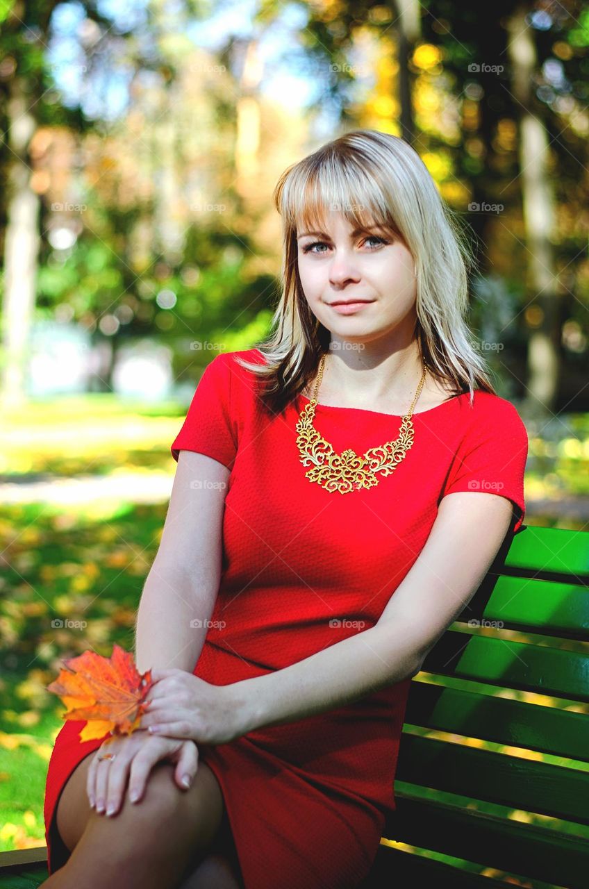 Portrait of beautiful young blonde woman, girl in red dress sitting at the bench in the autumn park.