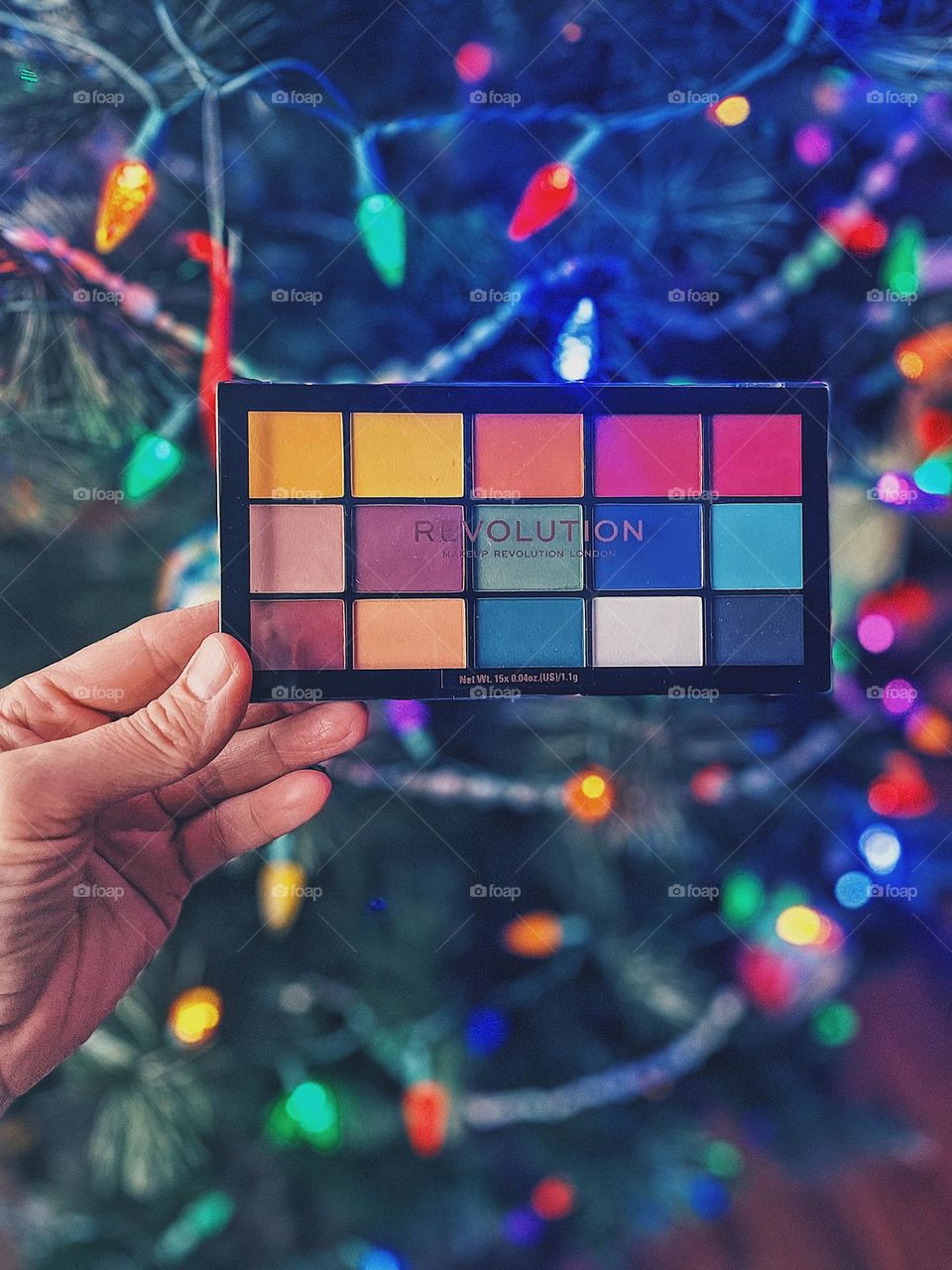 Woman holding eye shadow palette over Christmas tree, woman’s hand holding beauty product, Revolution makeup product, Revolution eye shadow palette, beauty products for a Christmas present, placing beauty products under the Christmas tree
