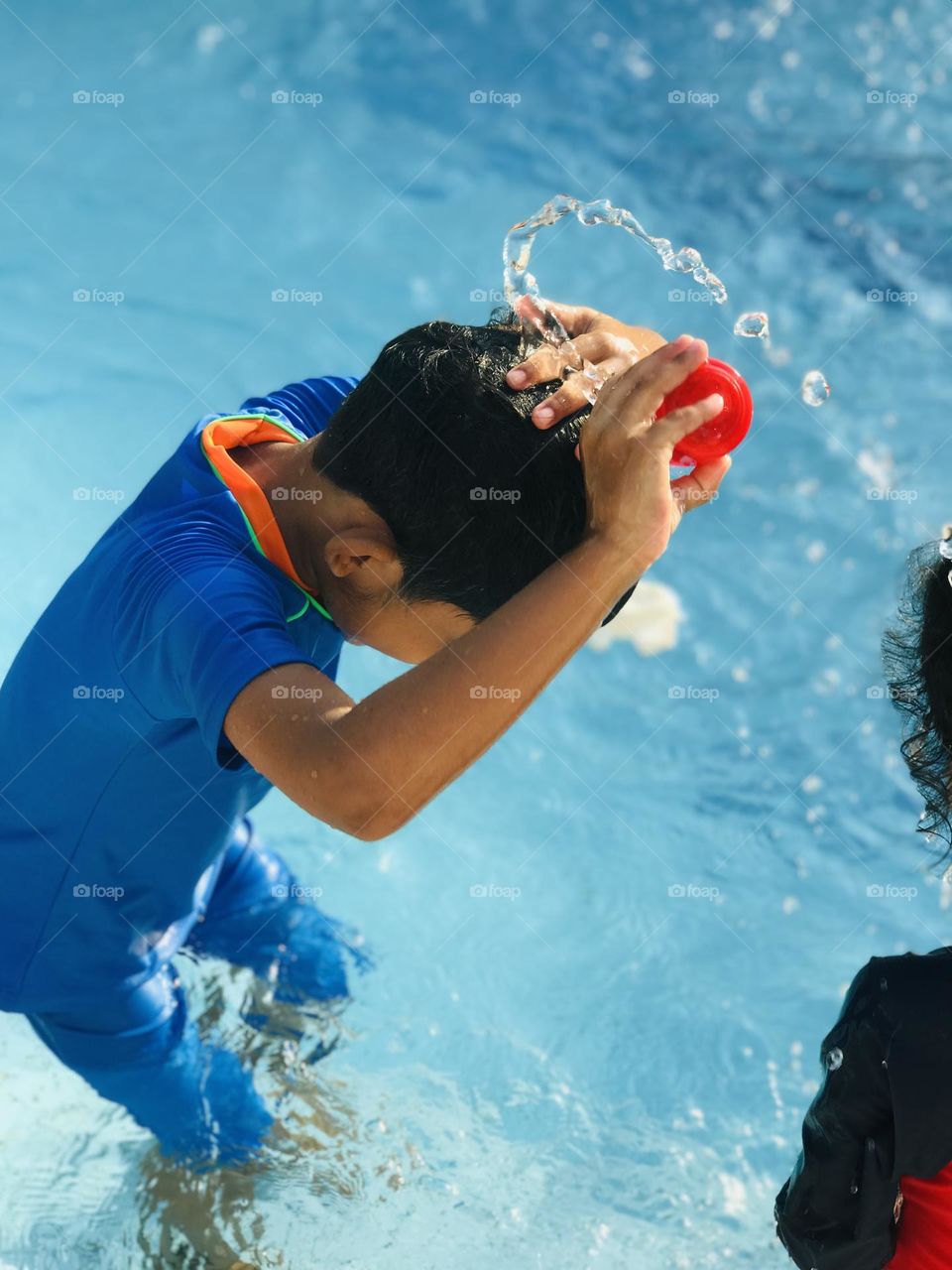 A funny boy playing with water by pouring water on his head at swimming pool in summer .